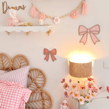 Load image into Gallery viewer, Children's bedroom with pink and white decor, including a bed with checkered blanket and wicker headboard, nightstand with lamp, and wall decorations.