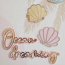 Load image into Gallery viewer, Decorative wall art featuring the phrase 'Ocean dreaming' in metallic script alongside three seashell shapes in pale blue, cream, and pink, arranged on a light beige background with a subtle palm leaf pattern.
