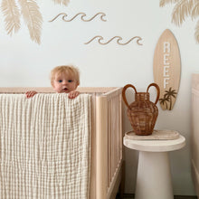 Load image into Gallery viewer, A baby nursery scene with a toddler peeking over a light wood crib draped with a cream check blanket. The room has tropical palm tree wall decals, a surfboard decoration, and a matching light wood dresser.
